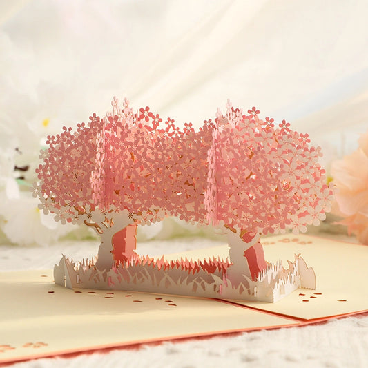 Close-up view of the 3D cherry blossom pop-up card with delicate pink paper flowers forming tree canopies, set against a blurred background of white and pink artificial blooms for a romantic aesthetic.