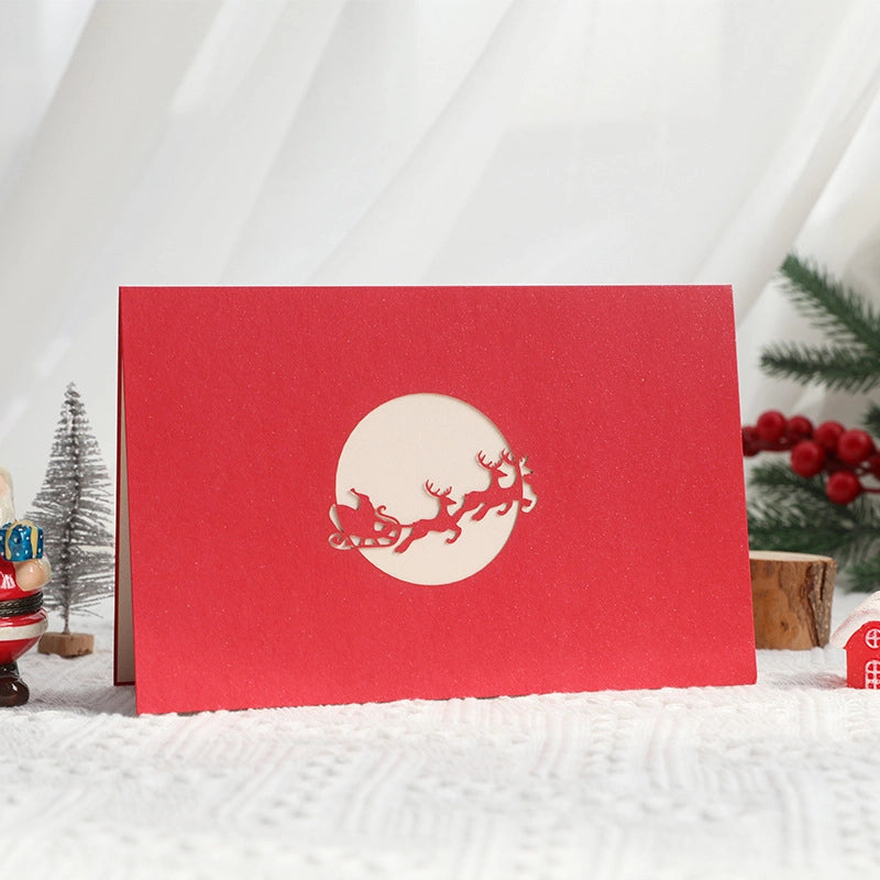 Red Christmas card cover with a laser-cut moon design revealing a silhouette of Santa's sleigh flying across the night sky.