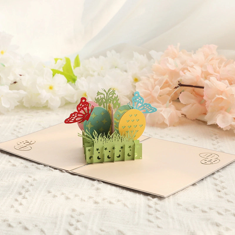 Open 3D Easter pop-up card featuring colorful eggs, red and blue butterflies, and green grass on a table with white and pink flowers in the background.