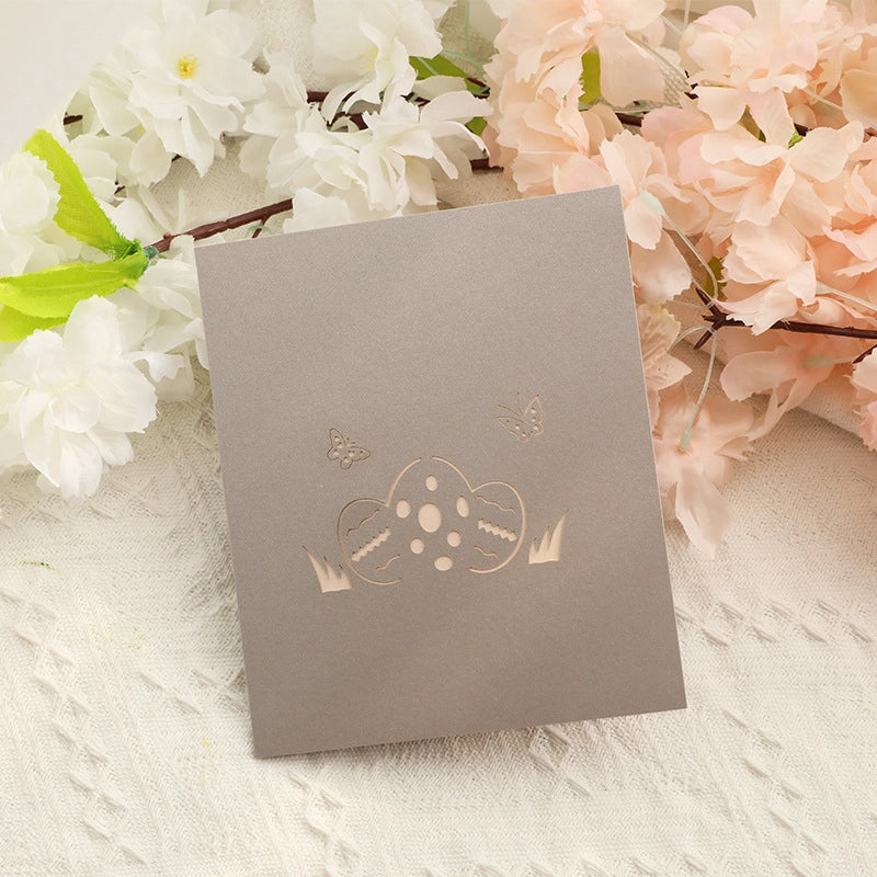 Closed beige Easter card cover with an elegant laser-cut design of an egg and butterflies, resting on a white knitted surface with flowers.