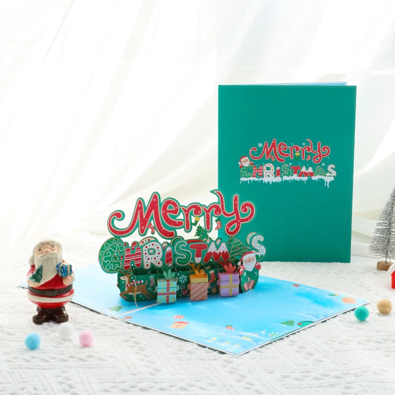 Open 3D Merry Christmas pop-up card showing red and green typography with gift boxes, next to the closed teal envelope.