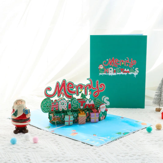Open 3D Merry Christmas pop-up card showing red and green typography with gift boxes, next to the closed teal envelope.