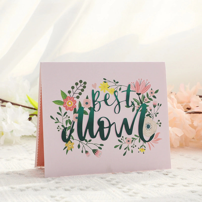 Pink greeting card cover featuring "Best Mom" calligraphy text surrounded by watercolor style flowers and leaves
