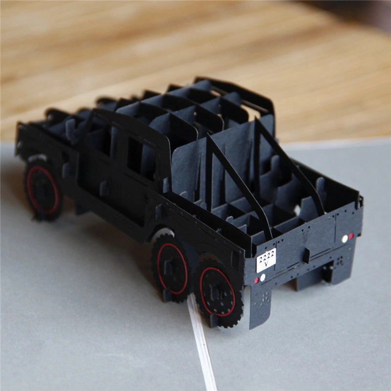 Side angle close-up of the black paper craft truck card, highlighting the red wheel details, license plate, and intricate layered construction on a wooden table.
