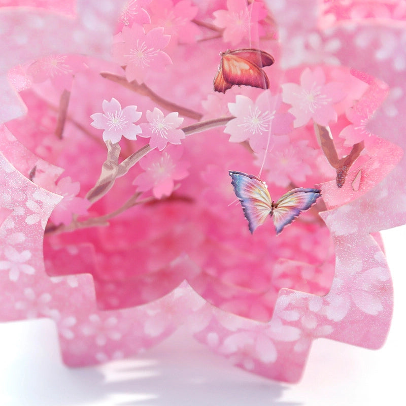 Close-up macro shot of the interior of a 3D pop-up card featuring detailed paper cut cherry blossoms and colorful butterflies hanging from branches
