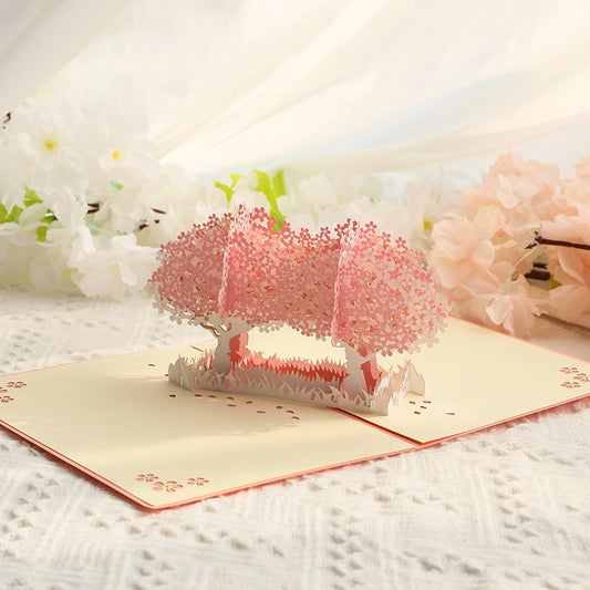 Romantic pink cherry blossom trees 3D pop-up card open on a white textured surface with soft floral background, showcasing intricate paper-cut sakura details and a dreamy spring atmosphere.