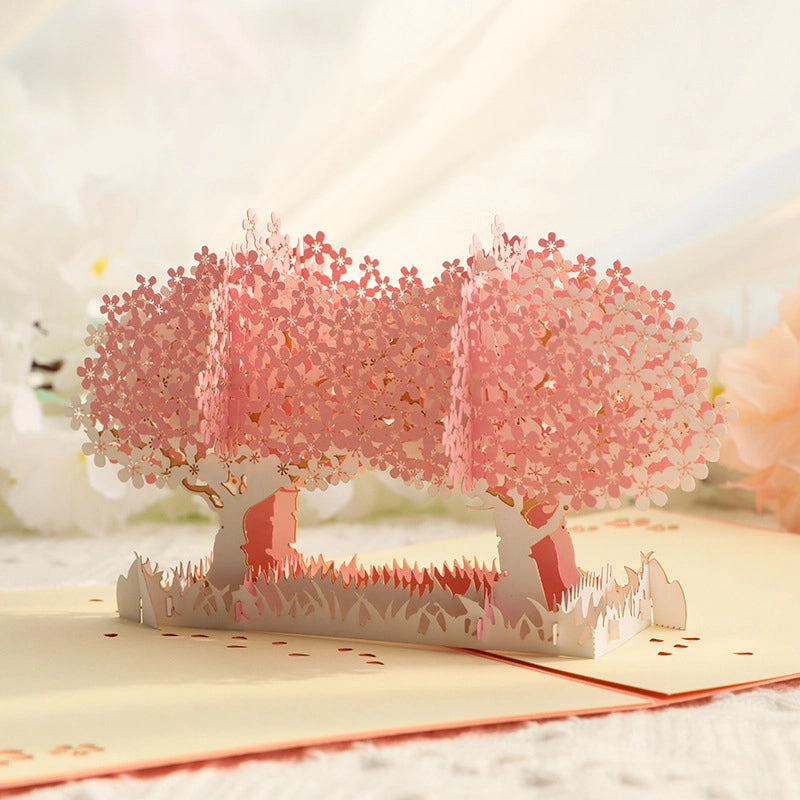 Close-up view of the 3D cherry blossom pop-up card with delicate pink paper flowers forming tree canopies, set against a blurred background of white and pink artificial blooms for a romantic aesthetic.