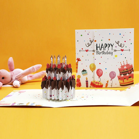 Opened 3D birthday greeting card showing a detailed paper cake sculpture with colorful balloons on the cover.
