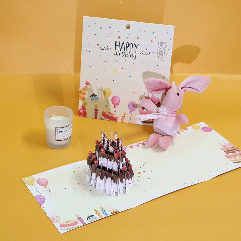 Lifestyle shot of the 3D birthday cake card displayed on a yellow table with a pink bunny toy and candle.
