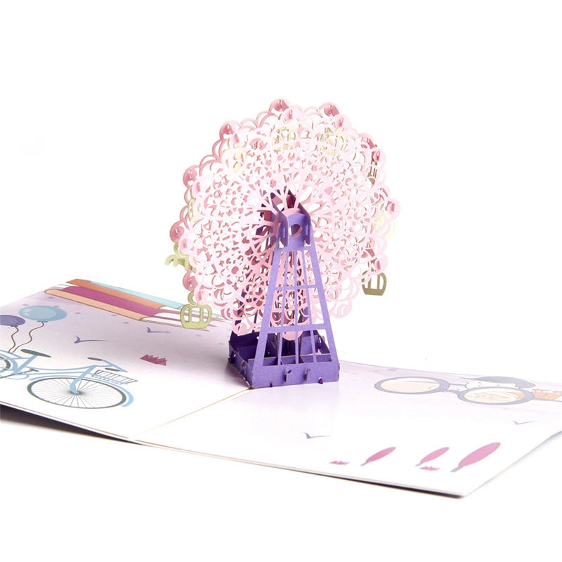 Close-up detail of intricate pink laser-cut Ferris wheel paper craft with green gondolas.
