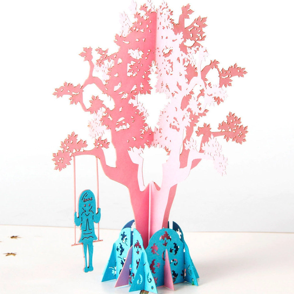 Side angle of open pop-up card displaying depth of pink tree structure and kraft paper envelope
