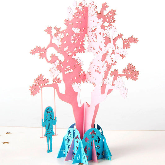 Side angle of open pop-up card displaying depth of pink tree structure and kraft paper envelope
