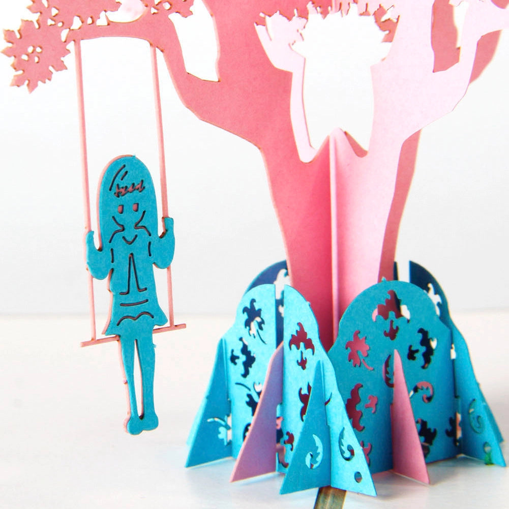 Close-up detail of blue paper cut girl on swing hanging from pink cherry blossom tree in 3D card
