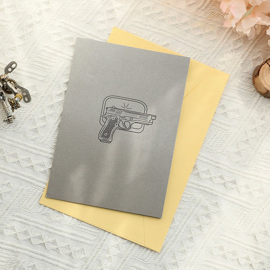 Grey greeting card cover with minimalist pistol line art design resting on a yellow envelope, creative gift stationery
