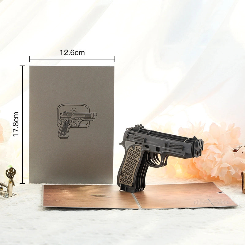 3D pop-up greeting card featuring a realistic paper pistol model standing on a wooden floor background, grey cover with gun outline, size 12.6x17.8cm
