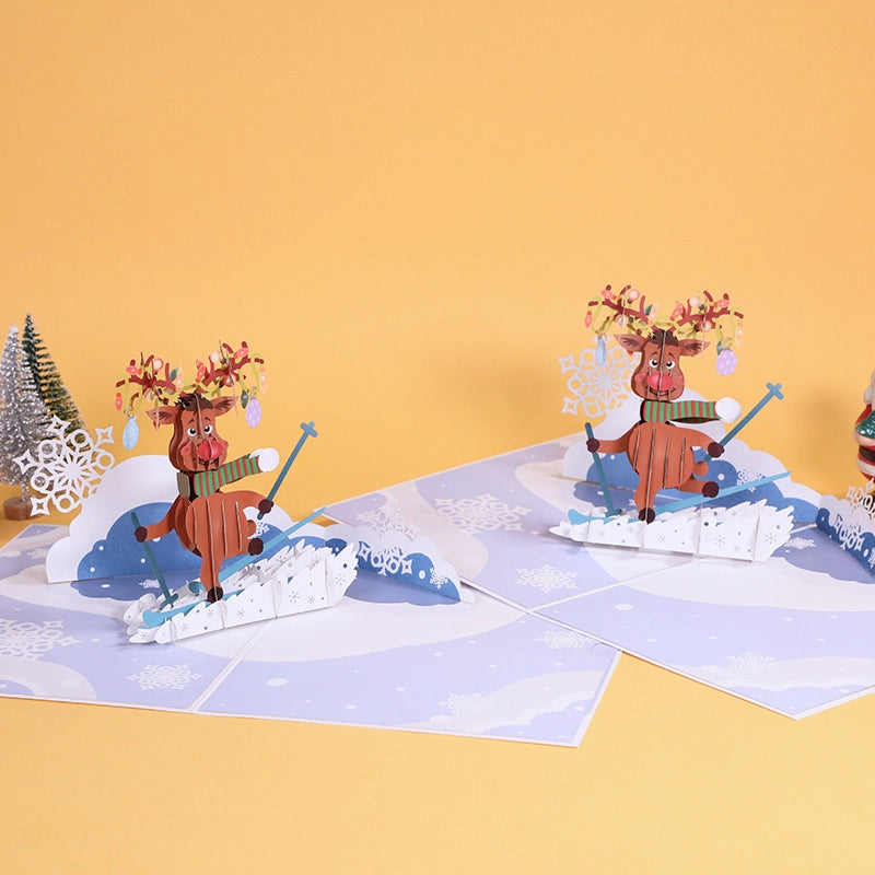 Skiing reindeer pop-up card displayed on a table with mini Christmas trees and Santa figurine.