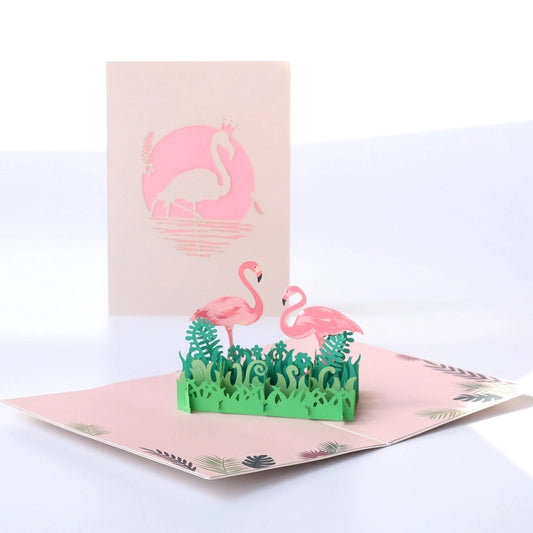 Open 3D pop-up card featuring two pink flamingos standing in green tropical grass, with a pink laser-cut cover showing a flamingo silhouette in the background.